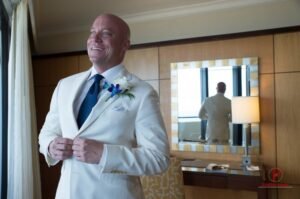 A man in white suit and blue tie standing next to a mirror.