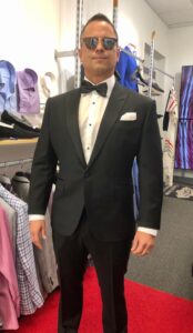 A man in a tuxedo standing next to a rack of suits.